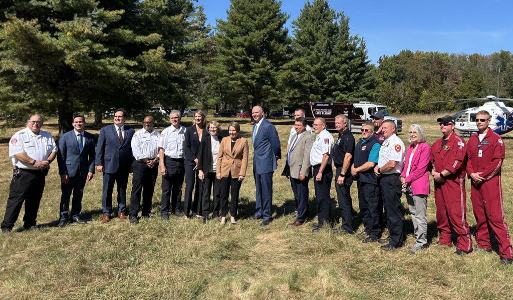 Group of people standing in a field with trees in the background, with a helicopter on the right and emergency response vehicles on the left.