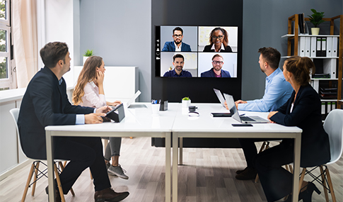 Four employees sit at table in conference room turned towards big monitor showing four other members for a meeting. 