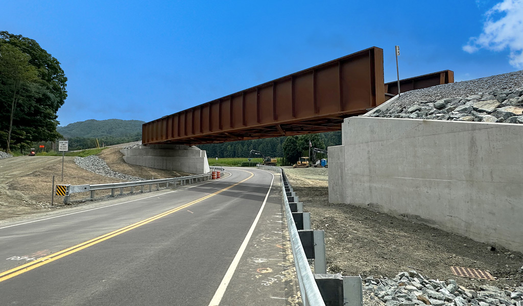 A newly constructed metal bridge over a road, with concrete supports and clear blue sky above.