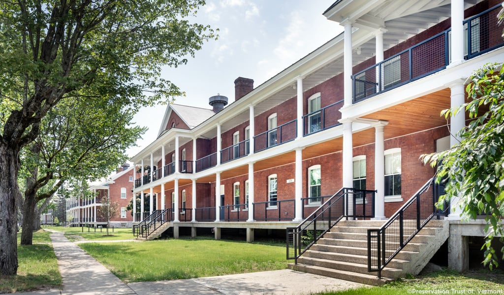 Front of a large brick building with white columns and metal railings fronted by trees, sidewalks, and a lawn
