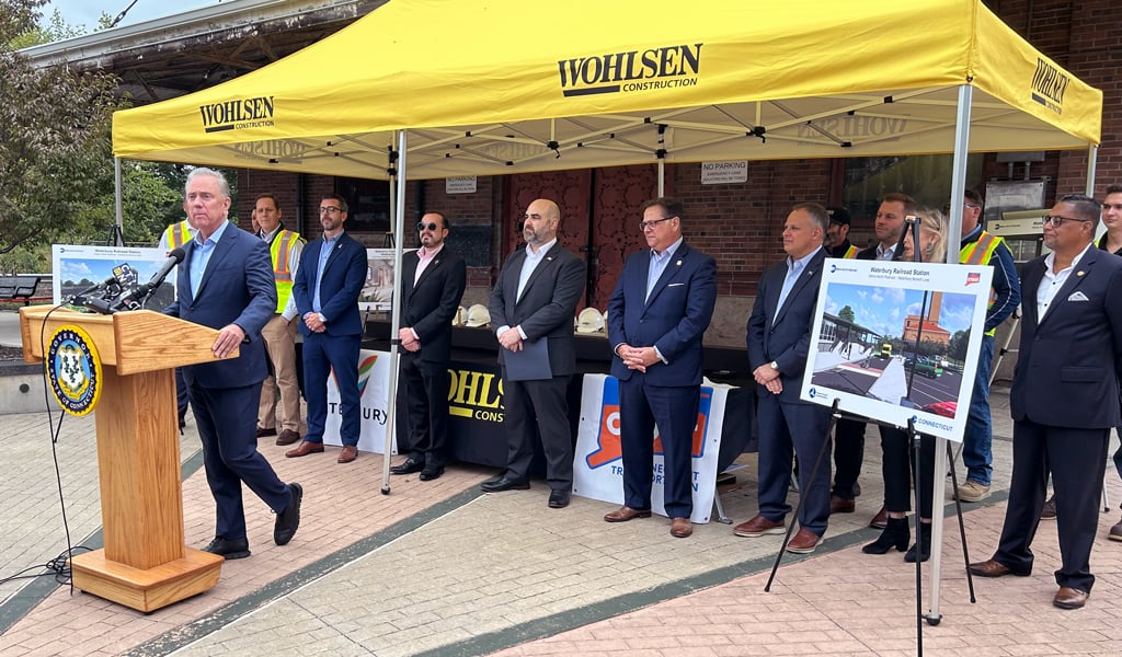 A man speaking at a podium with the Wohlsen Construction banner, surrounded by several individuals standing under a tent.