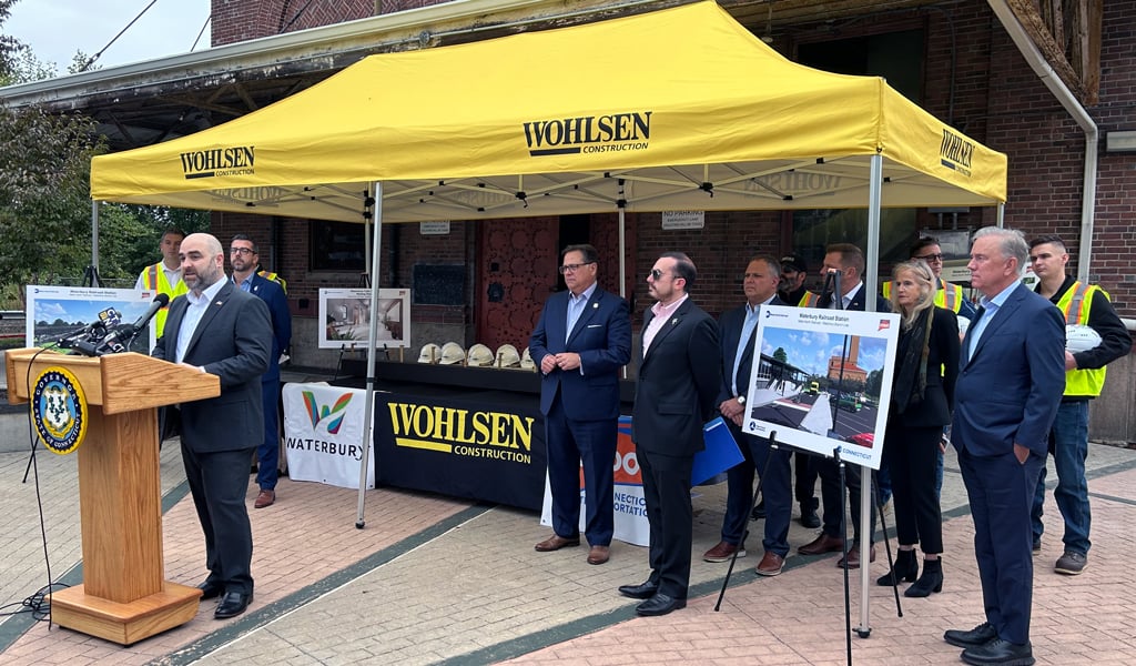 A man speaking at a podium with the Wohlsen Construction banner in front of a group of attendees at an outdoor event near a brick building.