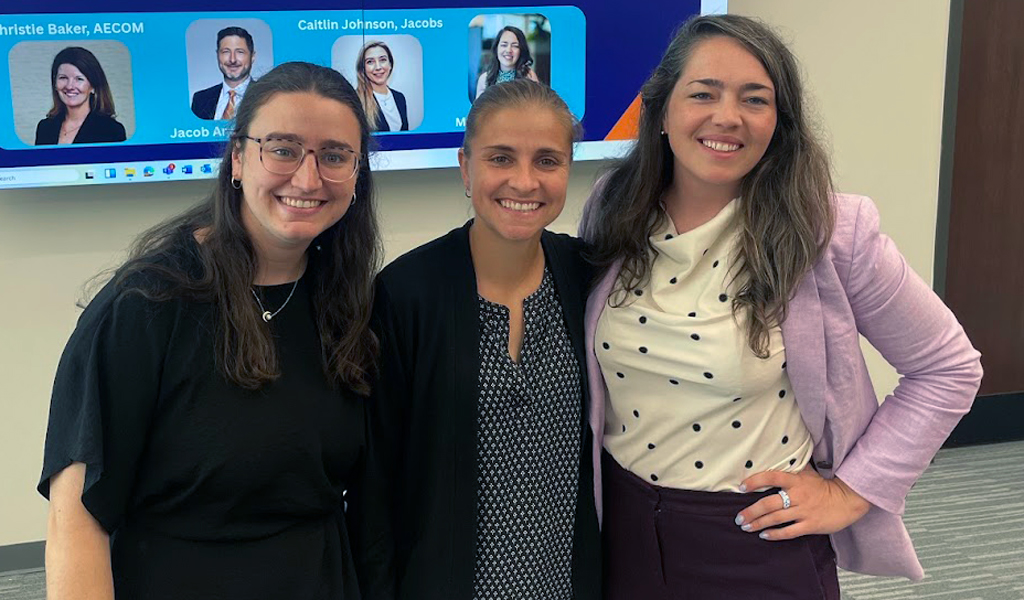 Three individuals smiling in front of a presentation screen at a WTS (Women's Transportation Seminar) event.