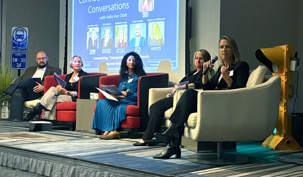 A panel of five people seated on stage facing the audience. Behind them, a projected slide shows the title "ConneCTed Conversations." Each panelist holds a microphone.  