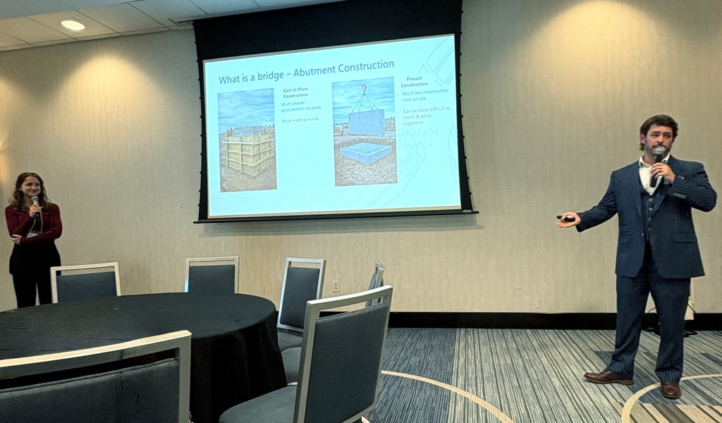 Two people in business attire are giving a presentation in a conference room. The presentation slide is titled "What is a bridge - Abutment Construction" and features images of construction materials and foundations.