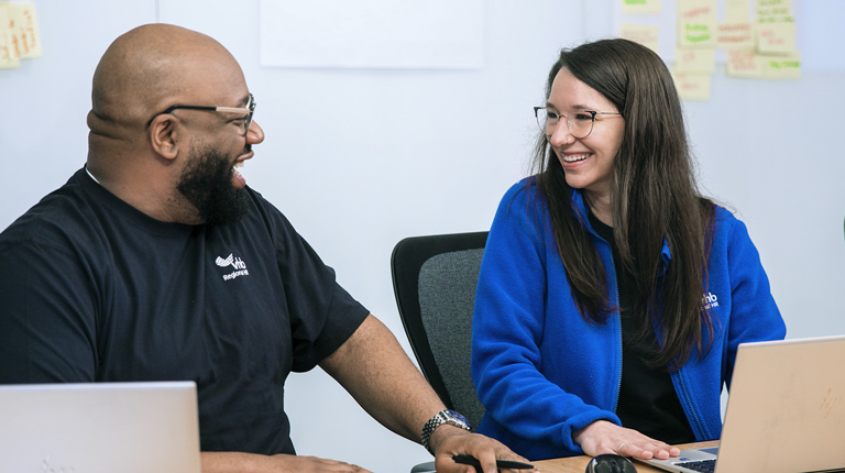 Two people sitting at a table with laptops, smiling at each other, with sticky notes on the wall behind them.