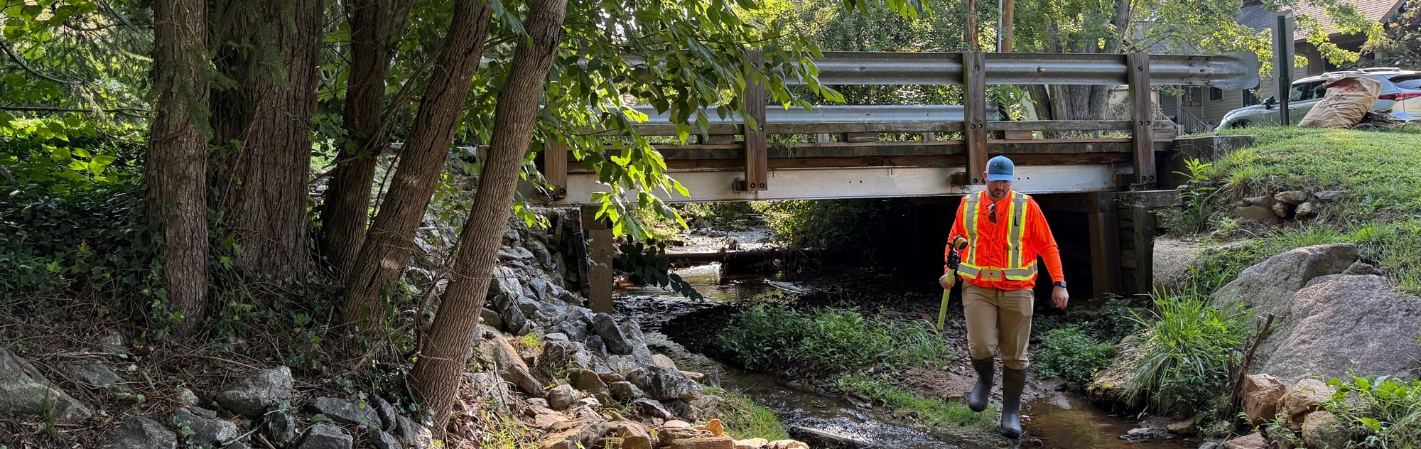 J.R. wading a shallow creek beneath a timber bridge during a hydraulic field investigation in Western North Carolina.