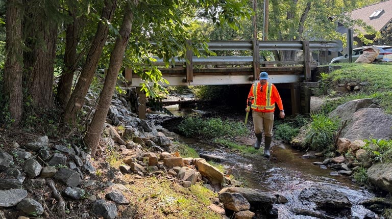 J.R. wading a shallow creek beneath a timber bridge during a hydraulic field investigation in Western North Carolina.