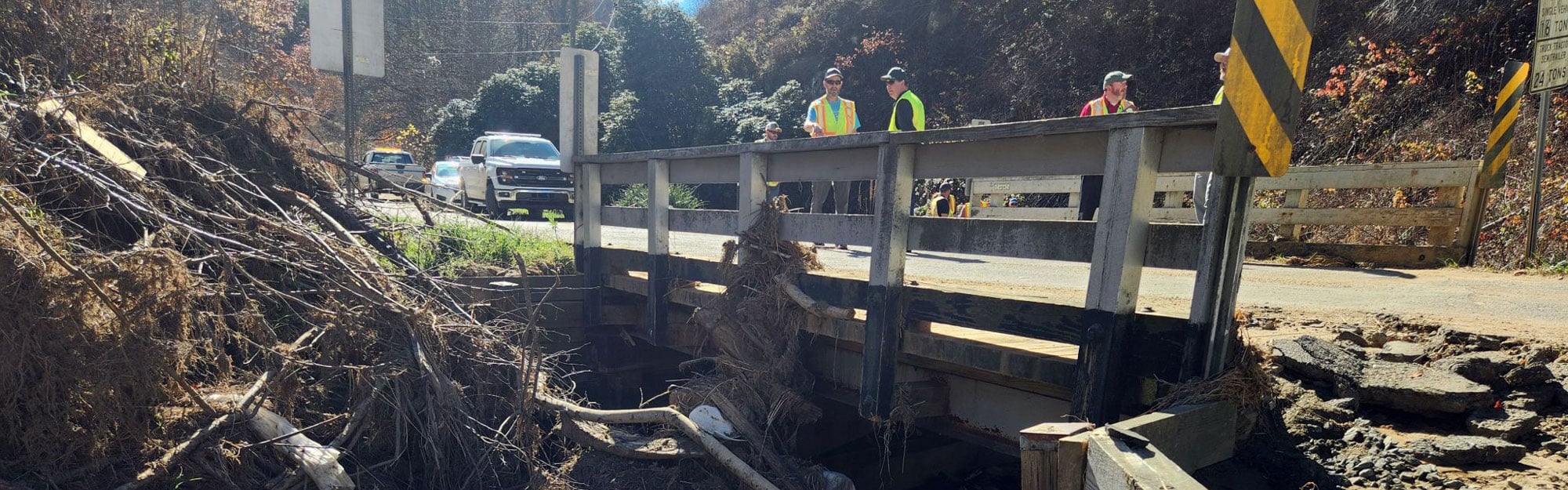 A small bridge spans a stream filled with debris and exposed sediment, people in high-visibility safety vests stand on the roadway above conducting an inspection.
