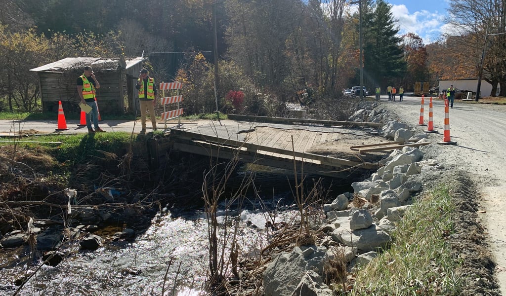 A two-lane rural bridge with a collapsed roadway spans a fast-moving, muddy river after severe flooding, leaving the road impassable while vehicles and utility lines are visible in the background.