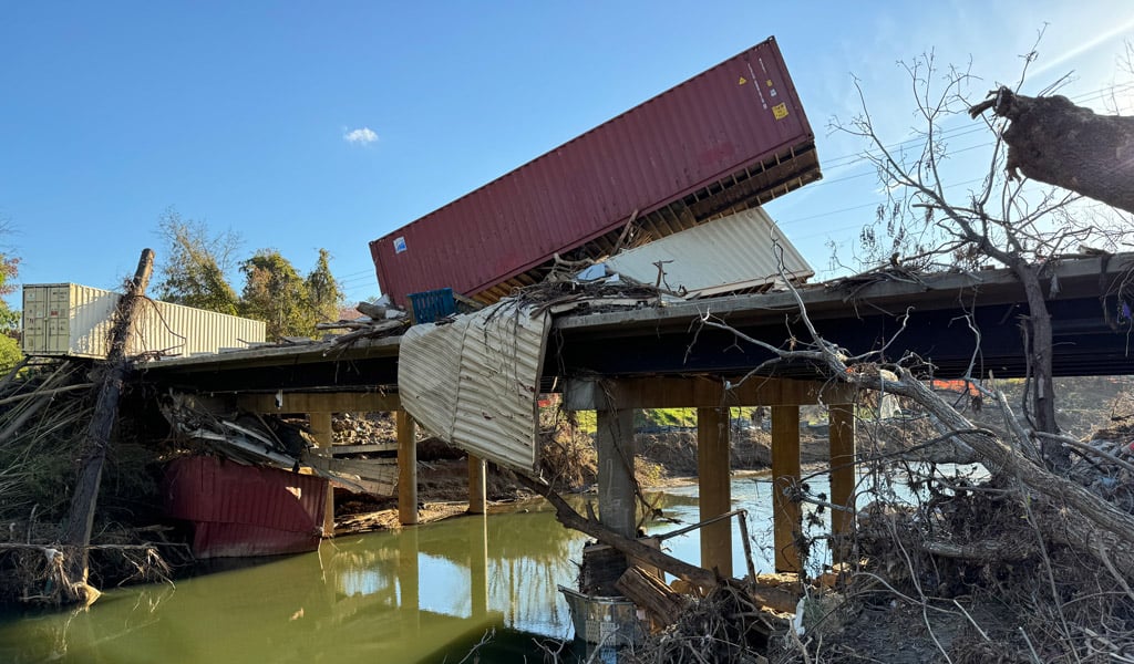 Damaged bridge spanning a river with multiple derailed shipping containers sitting on top; debris, twisted metal siding, and fallen tree limbs are caught on the structure&rsquo;s supports, showing significant storm-related damage.