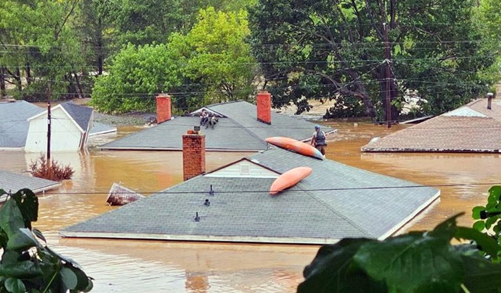 Single-story houses are partially submerged by muddy floodwater, with several people positioned on rooftops.