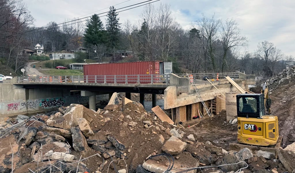 Bridge undergoing repair with a red shipping container positioned on the deck; exposed structure and active construction work are visible below, including scaffolding, ladders, and heavy equipment, with large rocks and soil piled near the riverbank.