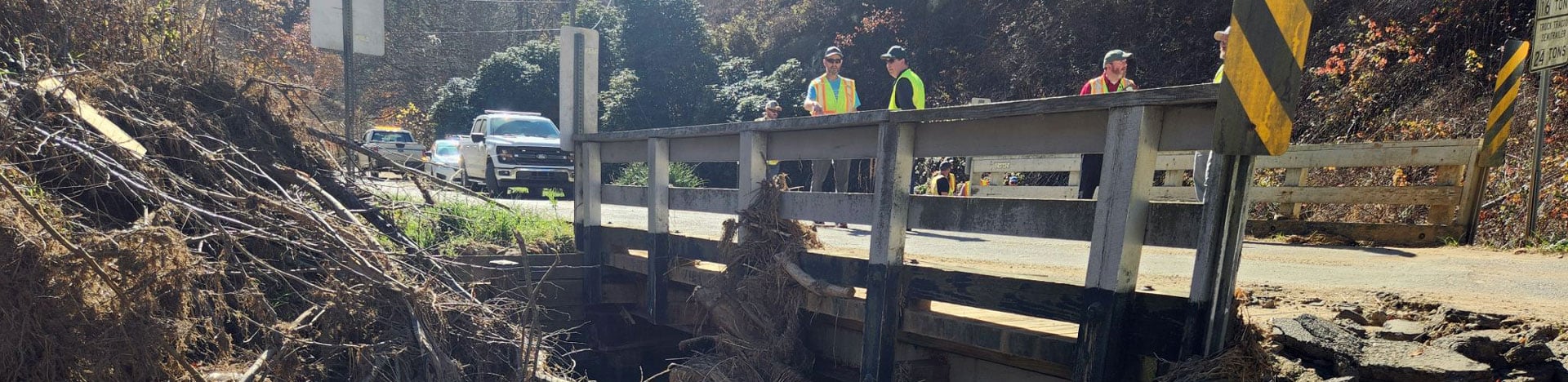 A small bridge spans a stream filled with debris and exposed sediment, people in high-visibility safety vests stand on the roadway above conducting an inspection.