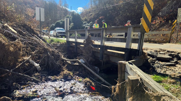 A small bridge spans a stream filled with debris and exposed sediment, people in high-visibility safety vests stand on the roadway above conducting an inspection.
