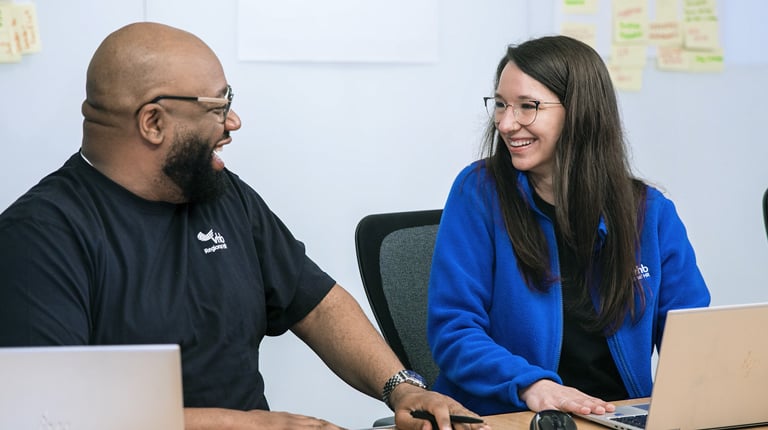 Two people sitting at a table with laptops, smiling at each other, with sticky notes on the wall behind them.