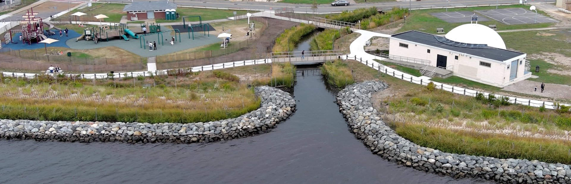 Waterfront park with playground, walking paths, nearby residential buildings, and a pier extending into water under a clear blue sky.