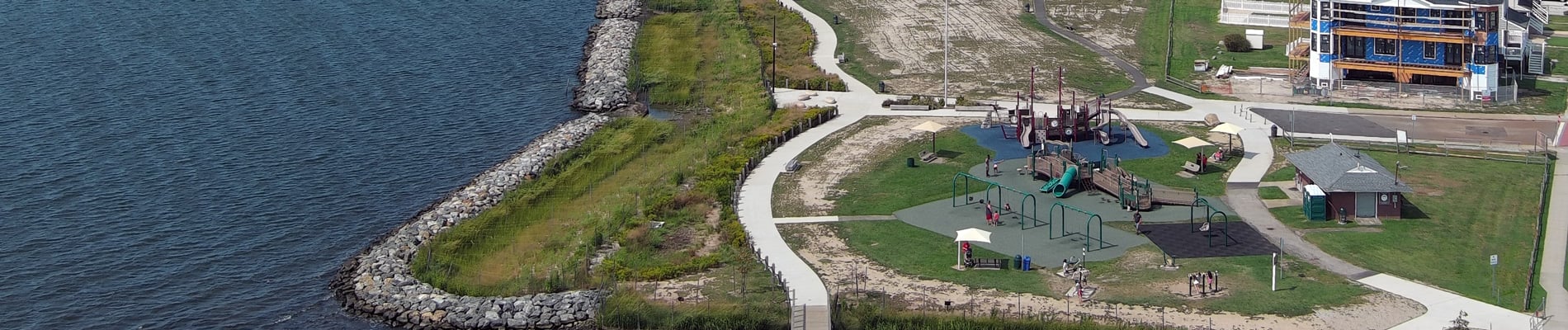 Waterfront park with playground, walking paths, nearby residential buildings, and a pier extending into water under a clear blue sky.