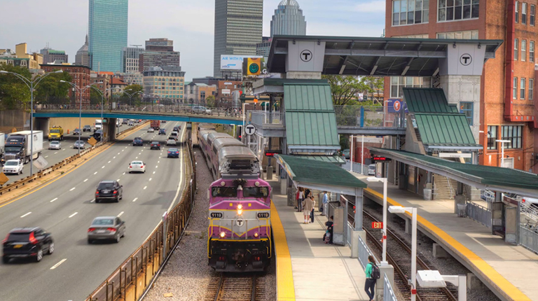 Commuters wait on a platform as a train pulls into the station, with the City of Boston in the background.