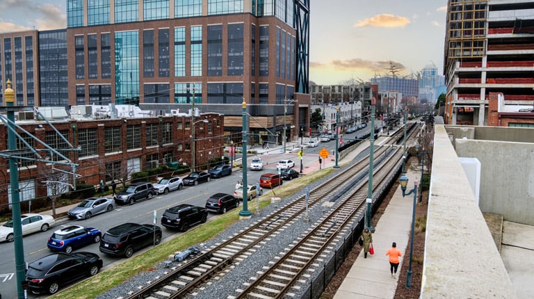 Urban redevelopment district with parallel light-rail tracks, busy roadway, modern office building, and construction site.